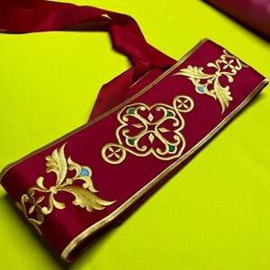 May include: A burgundy sash with gold embroidered floral and geometric designs. The embroidery includes teal and gold accents. The sash has a gold border and a long burgundy ribbon attached. The background is a bright yellow-green.