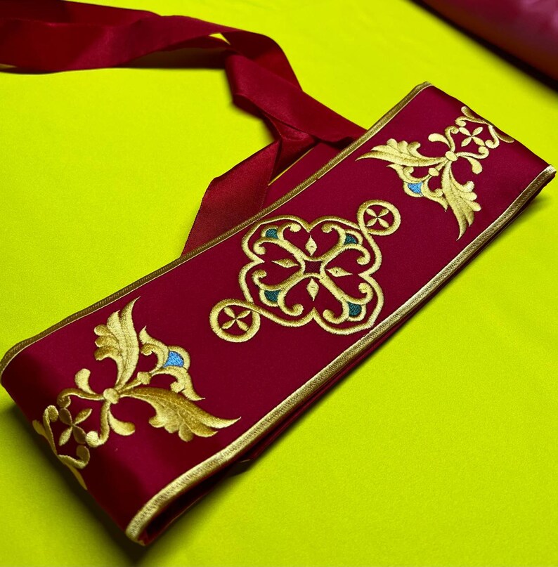 May include: A burgundy sash with gold embroidered floral and geometric designs. The embroidery includes teal and gold accents. The sash has a gold border and a long burgundy ribbon attached. The background is a bright yellow-green.