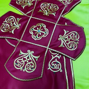 May include: Close-up of a burgundy textile with gold and green embroidered floral designs. The fabric is divided into square and curved sections, outlined with gold trim. The background is a bright green.