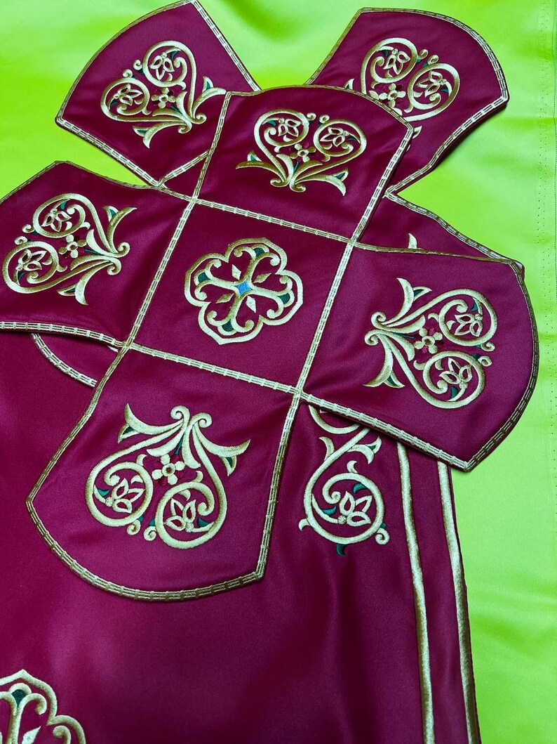 May include: Close-up of a burgundy textile with gold and green embroidered floral designs. The fabric is divided into square and curved sections, outlined with gold trim. The background is a bright green.