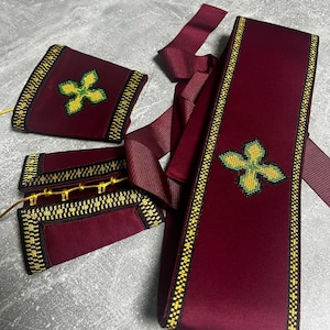 K&ouml;nnte beinhalten: Ein Set aus bordeauxroten Samt-Sch&auml;rpen mit goldenen und schwarzen Verzierungen. Jede Sch&auml;rpe ist mit einem gr&uuml;nen und gelben gestickten Kreuz verziert.