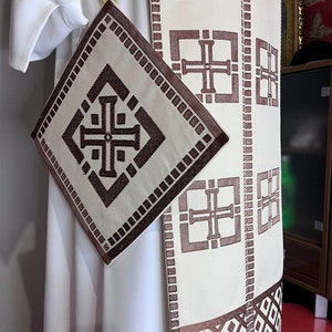 May include: A white fabric stole with a brown embroidered pattern of crosses and squares. The stole has a matching brown fabric pocket attached to it.