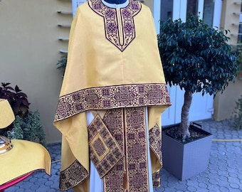 May include: A yellow liturgical vestment with intricate purple embroidery. The vestment has a long, flowing design and a decorative collar. The vestment is displayed on a mannequin in front of a building with a sign that reads "Благойст".