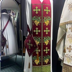 K&ouml;nnte beinhalten: Ein wei&szlig;es liturgisches Gewand mit einer burgunderfarbenen Stola, die mit gr&uuml;nen Weinreben und goldenen Kreuzen bestickt ist. Die Stola hat eine burgunderfarbene Tasche mit einem goldenen Kreuz.