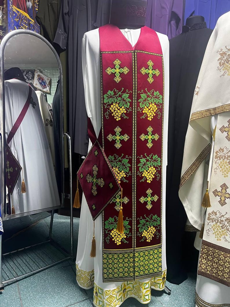 K&ouml;nnte beinhalten: Ein wei&szlig;es liturgisches Gewand mit einer burgunderfarbenen Stola, die mit gr&uuml;nen Weinreben und goldenen Kreuzen bestickt ist. Die Stola hat eine burgunderfarbene Tasche mit einem goldenen Kreuz.