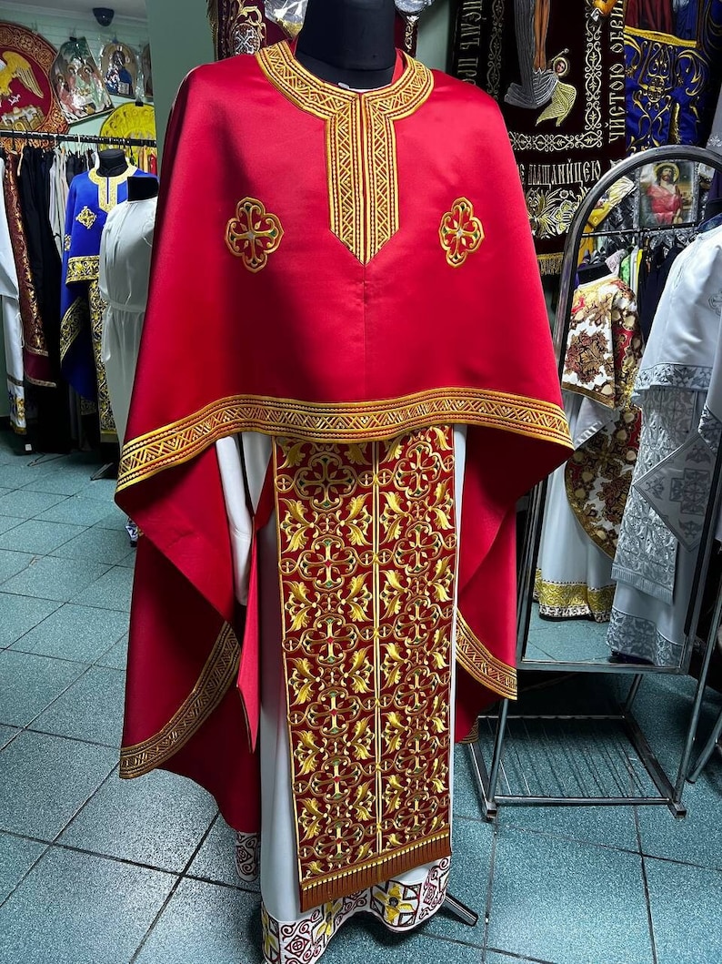 May include: A red liturgical vestment with gold embroidered details. The vestment features a cape-like design with intricate patterns around the neckline, hem, and central panel. The garment is displayed on a mannequin, showcasing its ornate design and craftsmanship.