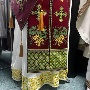 K&ouml;nnte beinhalten: Ein wei&szlig;es liturgisches Gewand mit goldenen und gr&uuml;nen Stickereien. Das Gewand hat ein rotes Feld mit einem Kreuz und Traubenranken-Motiven. Der Saum des Gewands ist gewellt und mit aufwendigen goldenen Stickereien verziert.