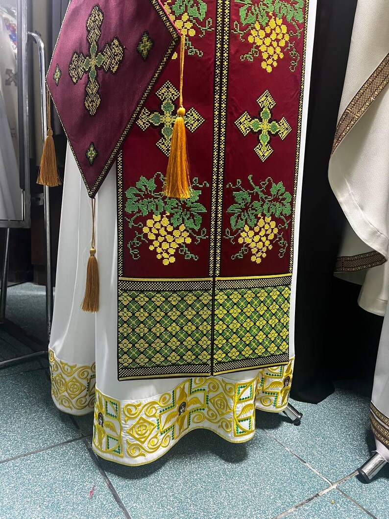 K&ouml;nnte beinhalten: Ein wei&szlig;es liturgisches Gewand mit goldenen und gr&uuml;nen Stickereien. Das Gewand hat ein rotes Feld mit einem Kreuz und Traubenranken-Motiven. Der Saum des Gewands ist gewellt und mit aufwendigen goldenen Stickereien verziert.