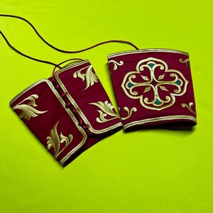 May include: Two burgundy-colored wrist cuffs with gold embroidery and trim. One cuff features a floral design, while the other has an ornate pattern. The cuffs are connected by a thin, dark cord, suggesting they are part of a larger garment or accessory.