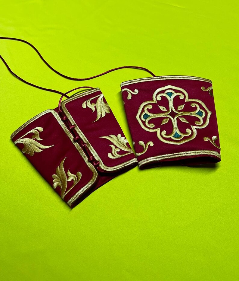 May include: Two burgundy-colored wrist cuffs with gold embroidery and trim. One cuff features a floral design, while the other has an ornate pattern. The cuffs are connected by a thin, dark cord, suggesting they are part of a larger garment or accessory.