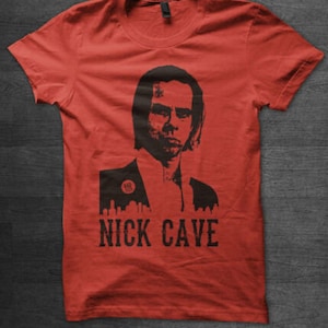 Può includere: Maglietta rossa con un'immagine in bianco e nero di Nick Cave. L'immagine mostra il viso e le spalle di Cave, e indossa un abito. Il testo "Nick Cave" è stampato sotto l'immagine.