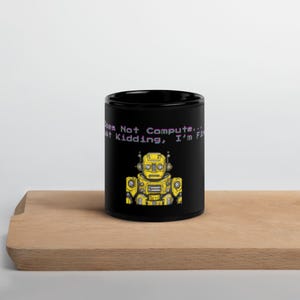 May include: Black ceramic mug featuring a yellow robot illustration with the text "Does Not Compute... Just Kidding, I'm Fine." The robot has a retro design. The mug is on a wooden surface.