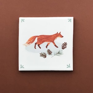 Könnte beinhalten: Eine weiße Keramikfliese mit einer handgemalten Illustration eines roten Fuchses, der durch einen Wald geht. Der Fuchs ist von Tannenzapfen und grünen Tannennadeln umgeben.