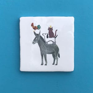 Könnte beinhalten: Quadratische Keramikfliese mit einer skurrilen Illustration eines Esels, eines Hundes, einer Katze mit Krone und eines Hahns. Die Tiere sind aufeinander gestapelt. Die Fliese ist weiß mit blauem Hintergrund.