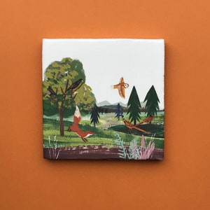 Könnte beinhalten: Quadratische Keramikfliese mit einer skurrilen Waldszene. Ein roter Fuchs springt über eine grüne Wiese, mit Vögeln auf einem Baum und einer fliegenden Eule. Das Kunstwerk zeigt Bäume, einen Pfad und Fasane.