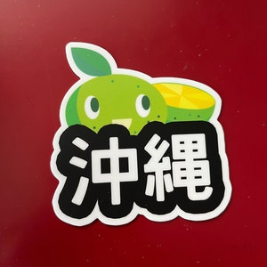 May include: A sticker featuring a cartoon lime with a smiling face and a slice of lime. The sticker has the Japanese characters for Okinawa in white on a black background, with a white border. The background is red.
