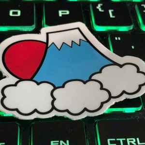 May include: A white sticker featuring a blue mountain with white snow and a red sun. The mountain is surrounded by white clouds. The sticker is placed on a black keyboard with green backlit keys.