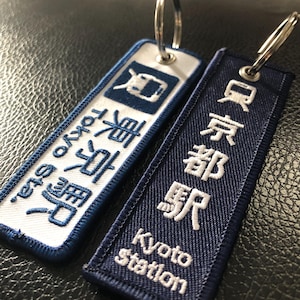 May include: Two rectangular keyrings. One is white with blue text saying "Tokyo" and a train icon. The other is navy blue with white text saying "Kyoto Station".