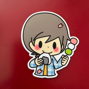Puede incluir: Una pegatina con un personaje de dibujos animados con cabello castaño, mejillas rosadas y una camisa a cuadros azul. El personaje sostiene una bola de arroz onigiri y un pincho de dango. Se incluyen acentos de flores de cerezo rosas.