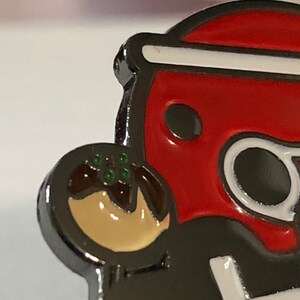 May include: A red and white enamel pin of a cartoon character holding a brown and beige takoyaki. The character has a black eye and a white outline.