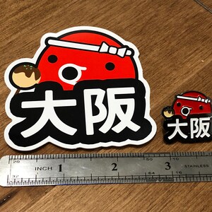 May include: Two Osaka themed pins, one large and one small. The larger pin is a red and white cartoon character with the text "Osaka" in black. The smaller pin is a similar design but smaller. Both pins are on a wooden surface with a ruler for scale.