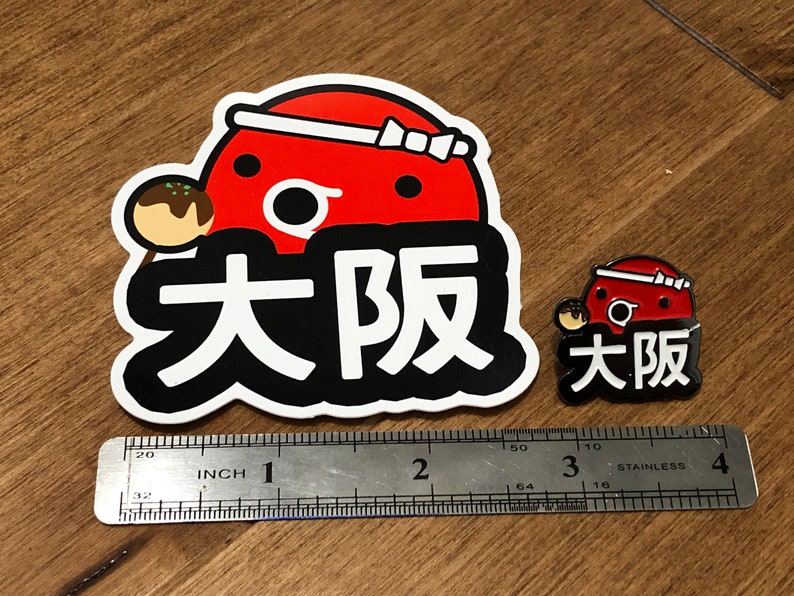 May include: Two Osaka themed pins, one large and one small. The larger pin is a red and white cartoon character with the text "Osaka" in black. The smaller pin is a similar design but smaller. Both pins are on a wooden surface with a ruler for scale.