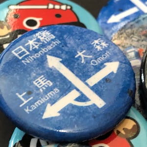 May include: Blue and white round button with Japanese text and arrows pointing in different directions. The text reads "Nihombashi", "Omori", and "Kamiuma".