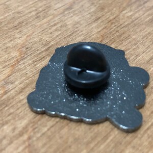 May include: The back of a silver metal pin with a black rubber backing. The pin features a detailed design with a glittery finish.
