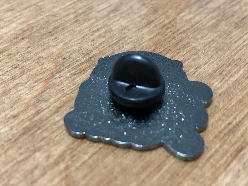 May include: The back of a silver metal pin with a black rubber backing. The pin features a detailed design with a glittery finish.