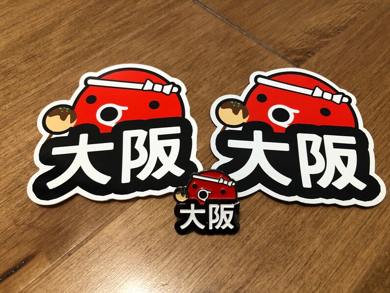 May include: Two red and white stickers with a cartoon octopus holding a takoyaki and the text "大阪" in black. A smaller red and white pin with the same design and text.