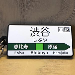 May include: A rectangular keychain featuring the Japanese characters for Shibuya station, with the English and Japanese names of nearby stations. The keychain has a white background with black text and green and black borders.