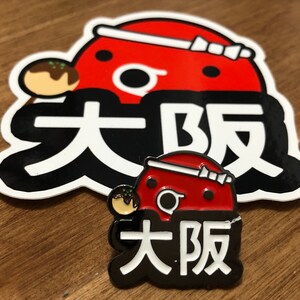May include: A red and black enamel pin with a cartoon character and the text "大阪" in white. The character is wearing a red hat with a white ribbon and holding a takoyaki.