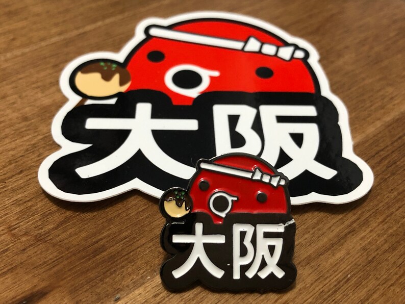 May include: A red and black enamel pin with a cartoon character and the text "大阪" in white. The character is wearing a red hat with a white ribbon and holding a takoyaki.