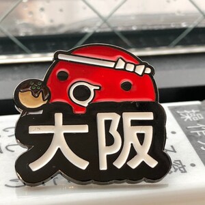 May include: A black and white enamel pin with a red and white cartoon character holding a takoyaki. The character has a white headband and the text "大阪" in white letters.