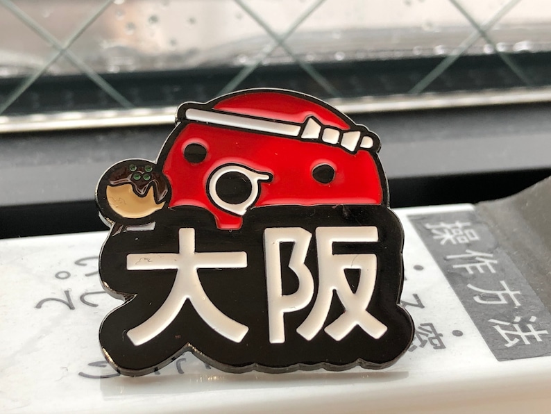 May include: A black and white enamel pin with a red and white cartoon character holding a takoyaki. The character has a white headband and the text "大阪" in white letters.