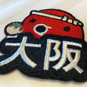 May include: A red and black embroidered patch with a cartoon character wearing a red hat and the Japanese text "大坂" in white.