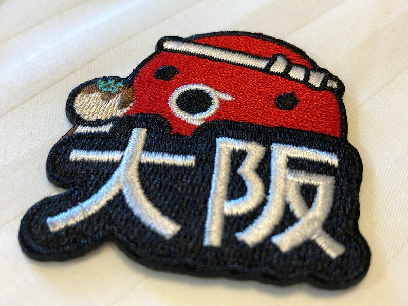 May include: A red and black embroidered patch with a cartoon character wearing a red hat and the Japanese text "大坂" in white.