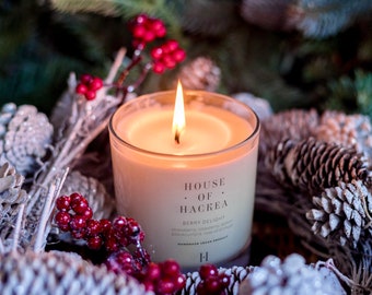 May include: A lit candle in a glass jar with a white label that reads "HOUSE OF HACKREA" and "BERRY DELIGHT" with a description of the scent: "strawberry, raspberry, lemon, blackcurrant, rose and musk". The candle is surrounded by a white and red holiday wreath.