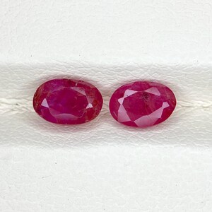 May include: Two oval-cut rubies with a deep red colour and faceted surfaces. Each gemstone displays a symmetrical shape with multiple triangular facets that reflect light, showcasing a translucent quality and a vibrant, saturated red hue.