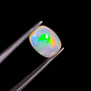 May include: A polished, oval opal gemstone held by tweezers against a black backdrop. The opal exhibits a play of colour, with flashes of green, orange, and blue within its translucent, milky white body. The gemstone is smooth and rounded.