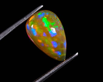 May include: A pear-shaped opal gemstone with a vibrant display of colors. The opal features a base of yellow and orange hues, with flashes of blue, green, and red. Held by tweezers against a black background, showcasing its unique iridescence.