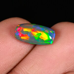 May include: A polished, oval opal gemstone. The opal exhibits a vibrant play-of-colour, with flashes of red, yellow, green, and blue. The gemstone is held between two fingers, showcasing its iridescent qualities against a dark background.
