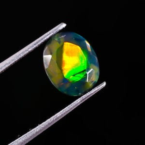 May include: An oval-cut gemstone held by tweezers. The gem displays a vibrant play of color, with flashes of yellow, green, and orange within a dark, translucent body. The facets catch the light, creating a sparkling effect against the black background.