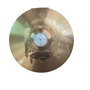May include: A round gold-toned metal compact case featuring an integrated analog clock face in the center. The surface is engraved with architectural motifs and decorative patterns. The clock has a white dial with black numerals and hands.