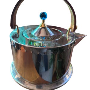 May include: A polished silver-tone metal kettle with a cylindrical body, a dark brown arched handle, and a conical lid topped with a small, round, bright blue finial. The kettle features a short spout and a reflective, smooth metallic surface.