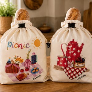 Peut inclure: Deux sacs à pain écrus à cordon de serrage avec des fermoirs noirs. Le sac de gauche présente une scène de pique-nique avec le texte "picnic" et un soleil. Le sac de droite montre une théière rouge à pois, une tasse et un gâteau.
