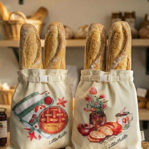 May include: Two beige bread bags, each holding baguettes. One bag has a pie graphic with "Autumn" and "Little Middle" text. The other features a floral design with bread and a mug. The bags have a white buckle closure.
