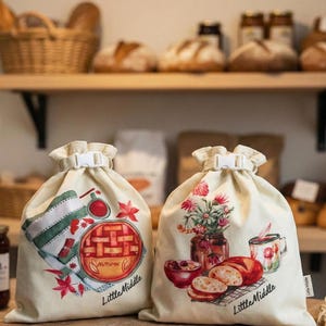 May include: Two cream-coloured drawstring bags with white plastic clasps. One bag features a pie graphic with the word "Autumn" and "Little Middle." The other bag has a floral arrangement and bread graphic with the same text.