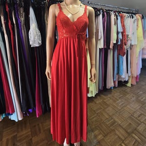 Puede incluir: Un camisón rojo largo hasta el suelo sobre un maniquí. El camisón presenta un corpiño de encaje con tirantes finos y un lazo en la cintura. Un collar de perlas adorna el cuello del maniquí. Otras prendas cuelgan en percheros en el fondo.