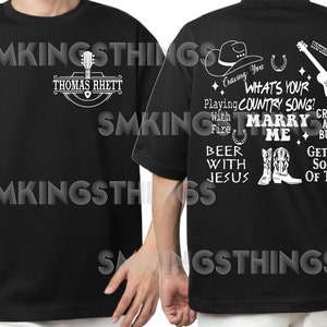 May include: Black t-shirt with a white graphic design featuring a cowboy hat, horseshoe, guitar, boots, and text that reads "Craving You, What's Your Country Song? Playing With Fire, Marry Me, Beer With Jesus, Crash And Burn, Get Me Some Of That".